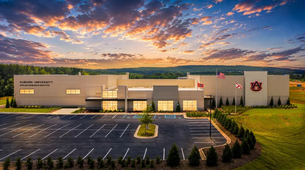 A modern building labeled "Auburn University Applied Research Institute" is surrounded by a parking lot and green landscaping under a partly cloudy sky.