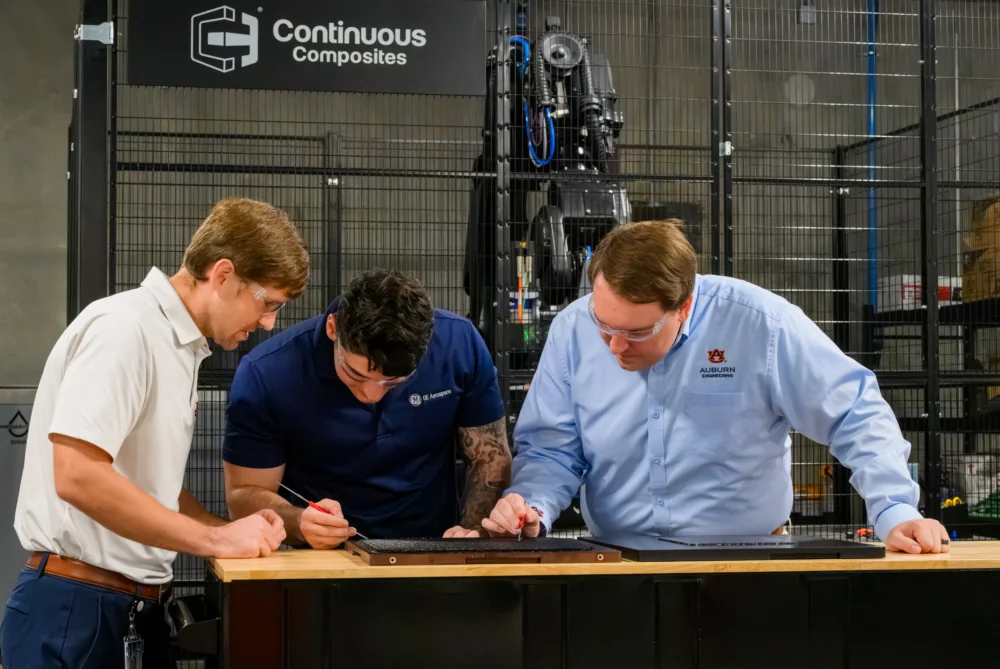 Three individuals collaborate on a workbench in a manufacturing facility, examining and adjusting components near a large machine.