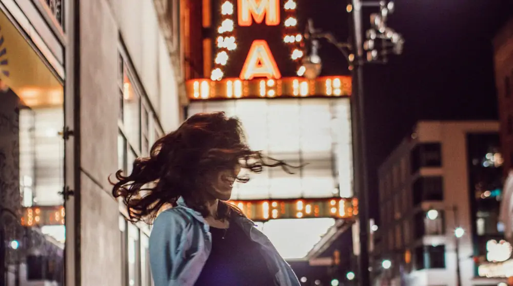 A woman joyfully spinning in front of the brightly lit Alabama Theatre marquee.