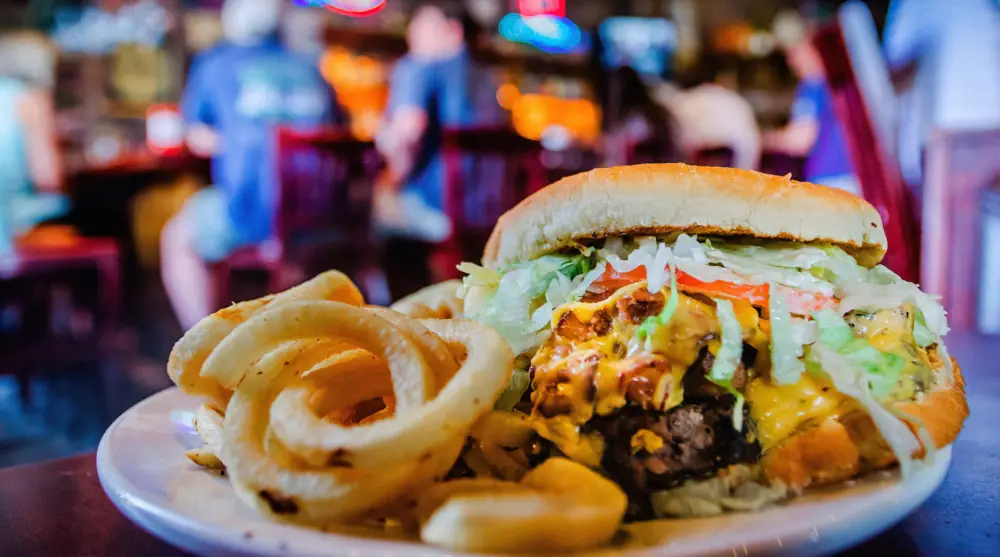 A delicious hamburger plate, dripping with cheese and sauce, with fries in sharp focus with a pub patrons sitting on stools in soft focus in the background.