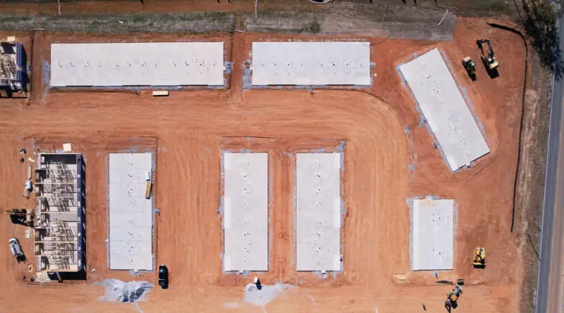 Aerial view of a commercial construction site in Alabama, showing concrete foundations and framing underway on red clay soil.