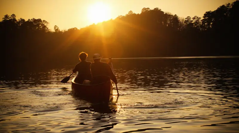 Two people paddling a canoe across a placid lake as the sun sets behind the tree line.