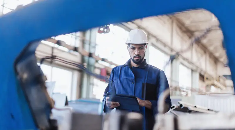 A man wearing a hard hat using a tablet seen through an opening of industrial equipment.