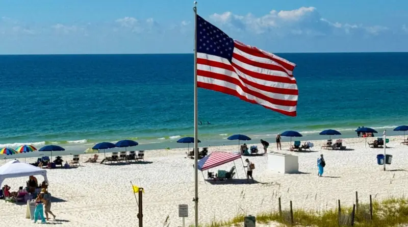 American flag waving on a pole above a crowded white-sand beach with turquoise water, beach umbrellas, and chairs on a sunny day.
